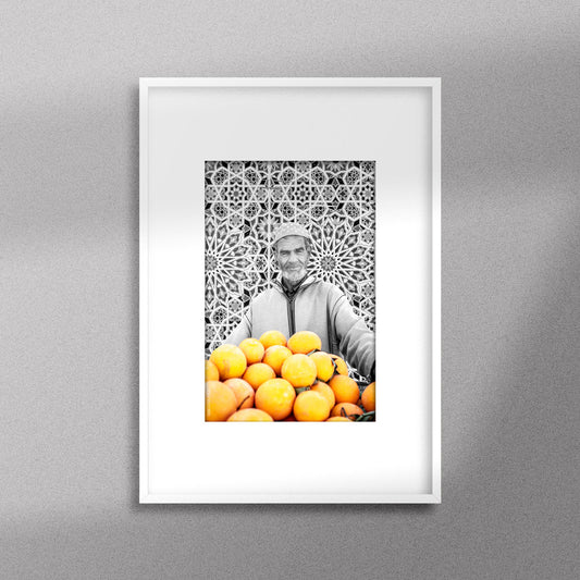 Tableau décoratif en noir et blanc d'un vieux marchand d'oranges très souriant, encadré dans un cadre blanc - Format A4.